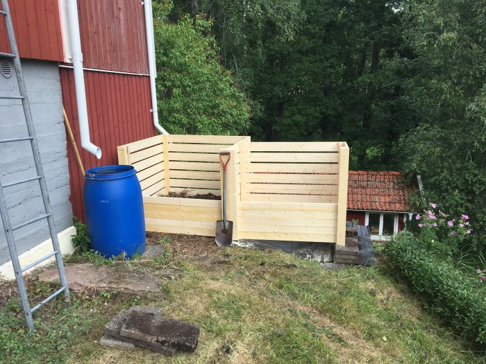 Två halvfärdiga byggda träkomposter intill en röd stuga och en blå tunna.