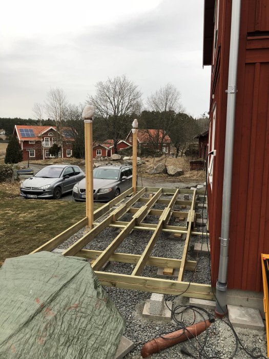 Ramverk och lagd trall för byggprojekt bredvid ett rött hus, med två stolpar framför och bilar i bakgrunden.