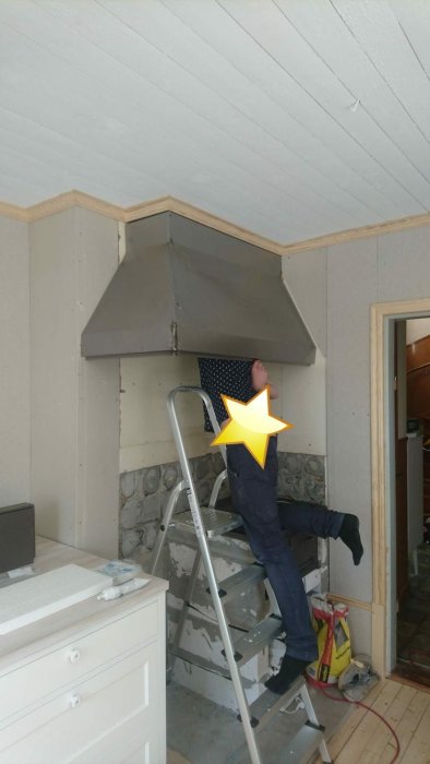 Monteringsarbete av grå kåpa ovanför spisplats med en person på en stege och köksinredning i förgrunden.