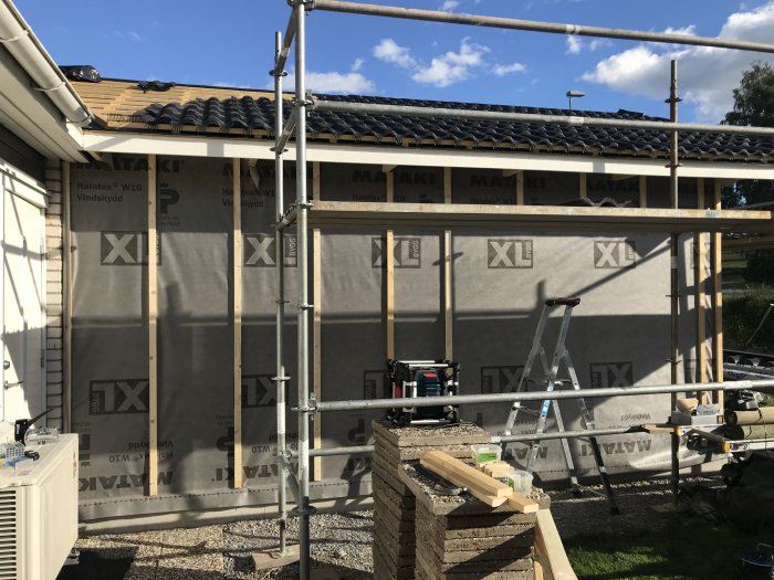 Husvägg med färdigreglad panelvägg och uppsatt vindskydd, byggmaterial och stege i förgrunden.