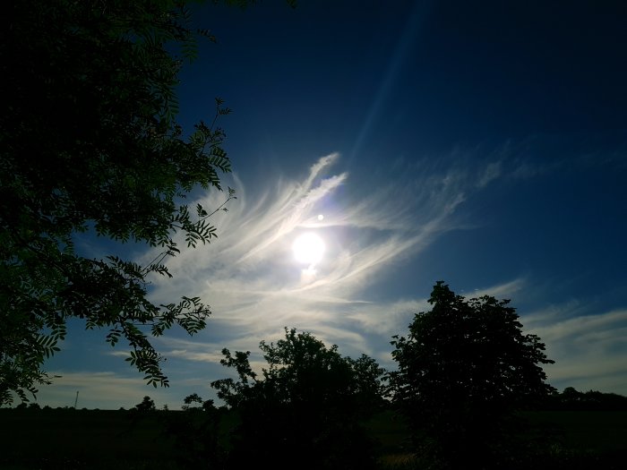 Sol genom molnstrimmor ovanför landskap med trädram i silhuett.