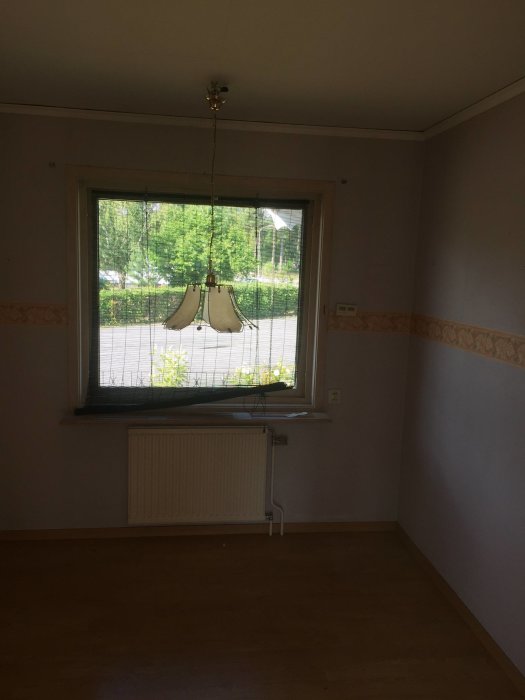 Äldre rumsinteriör med fönster, radiator, beige väggar och parkettgolv före renovering.