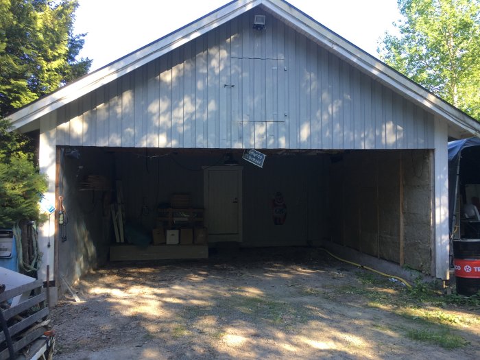 Öppen carport med tre väggar och diverse byggmaterial och verktyg inuti i förberedelse för ombyggnad till garage.