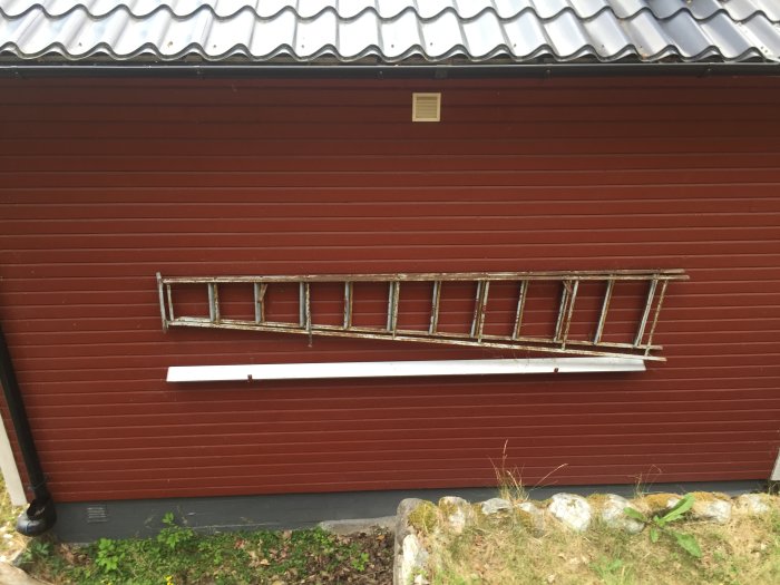 Rödmålad stugvägg med en vit stege lutad mot den och tecken på färg som släpper från panelen.