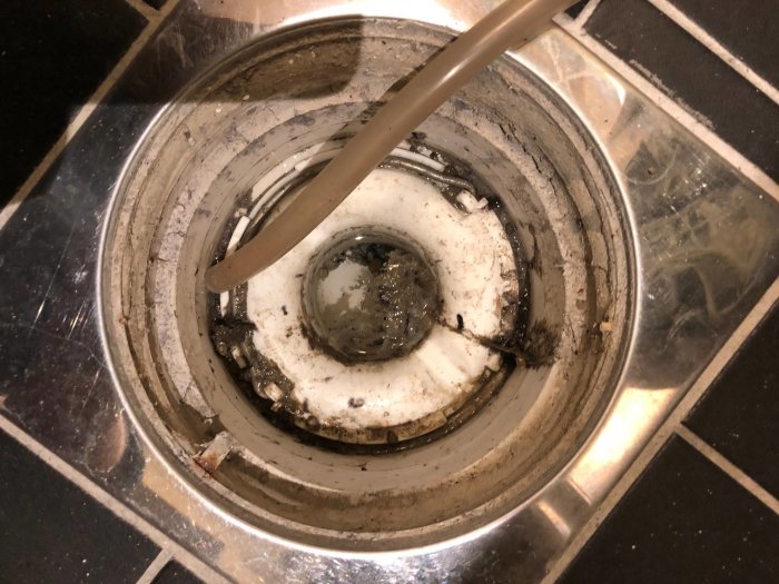 En sliten golvbrunn med en slang som ligger i för avfuktning, synligt smuts och skador.