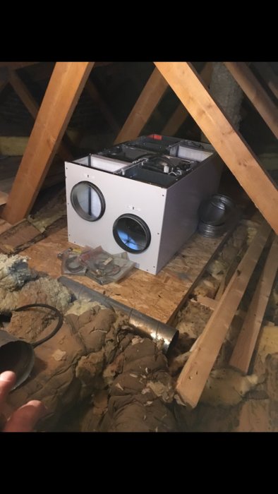 Ventilationsaggregat på en arbetsplattform i ett vindutrymme med OSB-skivor och isolering.