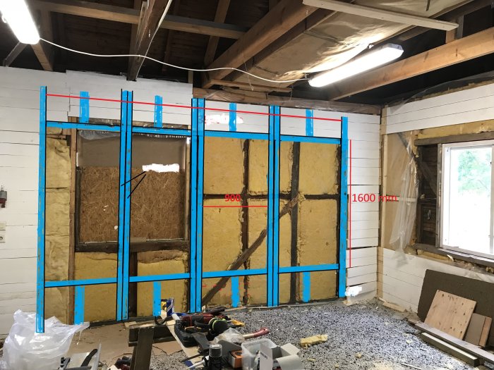 Skiss med blå markeringar för reglar och måttangivelser på en vägg under renovering, isolering synlig, byggmaterial utspridda.