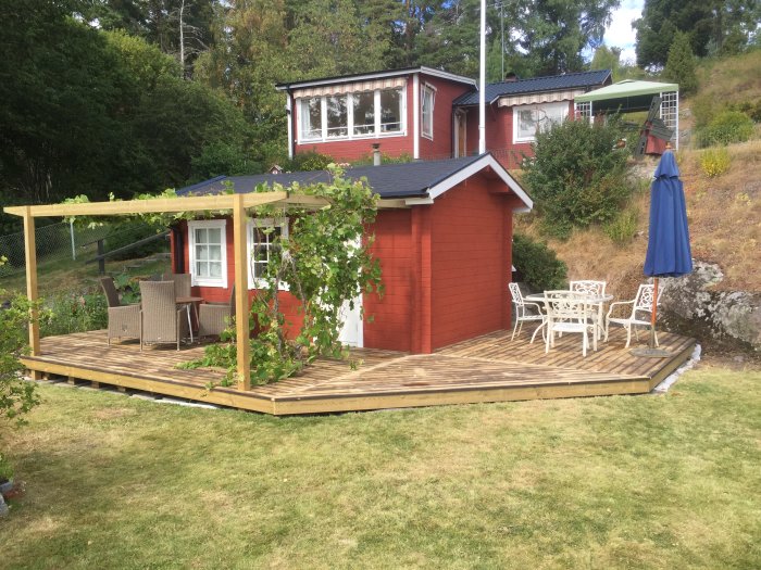 Altan byggd av återanvänt virke, upp-ned brädor, vid röd stuga med trädgårdsmöbler och pergola.