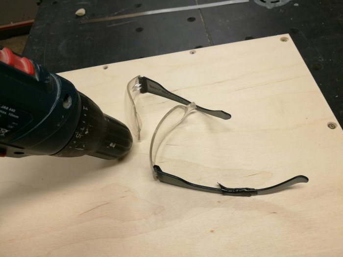 Bruten skyddsglasögon nära en skruvdragare på ett träbord, skalmen lagad med svart eltejp.