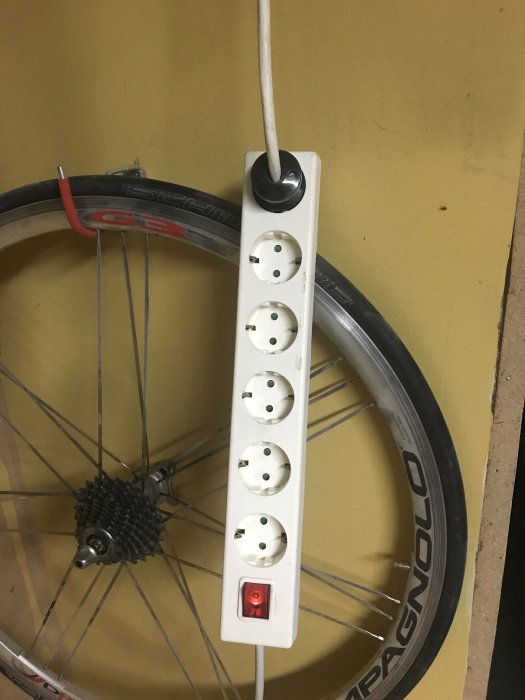 Vit grenuttag med stickpropp kopplad till gammal kabel, upphängd på cykelhjul mot gul vägg.