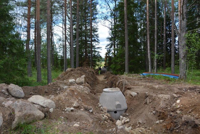 Bild av en grävd avloppsgröp i skog med betongbrunn och blå avloppsrör i förgrunden, grävmaskin och arbetare i bakgrunden.