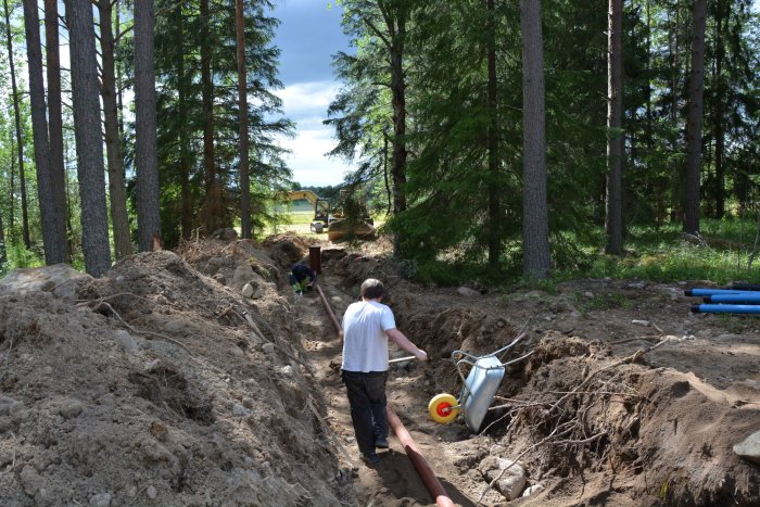 Personer arbetar med avloppsrören i en grävd dikesränna i skogsområde, med byggmaterial nära till hands.