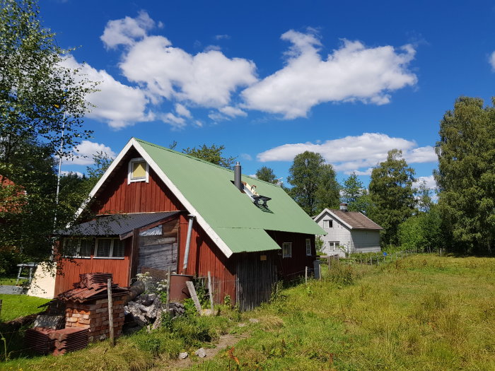 Röd trästuga med ny skorsten och grönt tak, väntande på plåtarbete, lantlig omgivning.