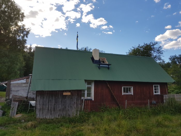 Ny skorsten installerad på taket till ett rött hus med grönt plåttak, omgivet av natur.