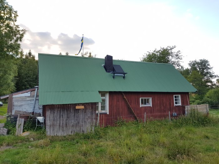 Rött trähus med ett nyinstallerat svart skorsten genom grönt plåttak, med svenska flaggan i bakgrunden.