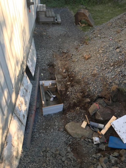 Upgrävning vid husvägg för att exponera och gjuta brunnsdel, med synliga rör och byggmaterial.