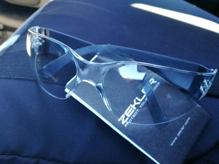 Skyddsglasögon från Zekler ligger på en mörk tygyta med texten "Protect Your Eyes" synlig.
