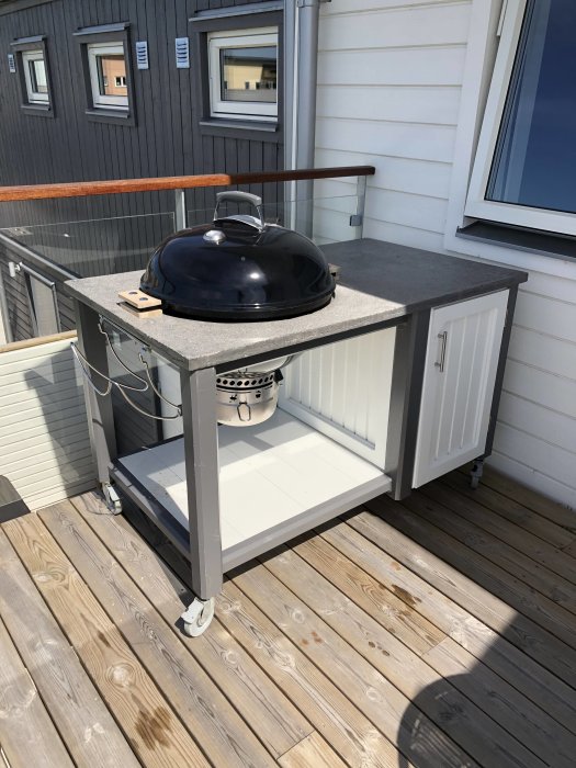 Handgjord grillbänk av reglar och plywood med en betongskiva och svart klotgrill på en träterrass.