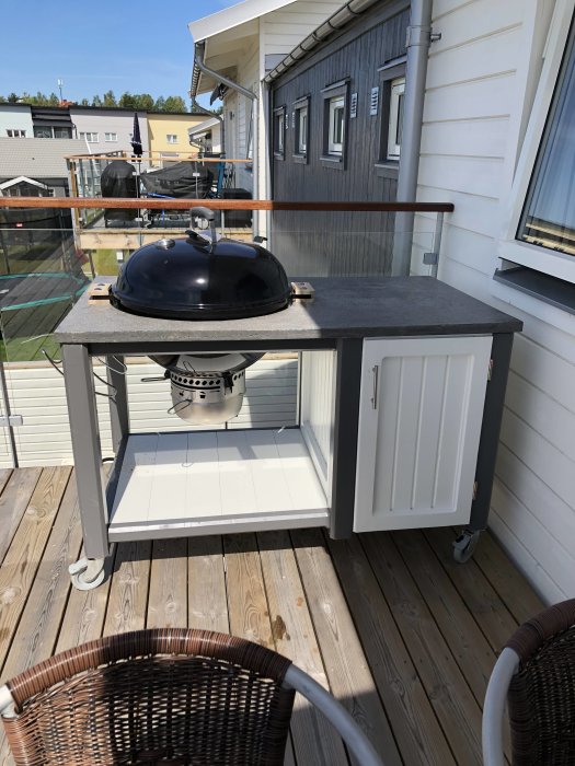 Hembyggd grillbänk med betongskiva och svart grillkupol på en träaltan bredvid en rottingstol, gjord av reglar och plywood.