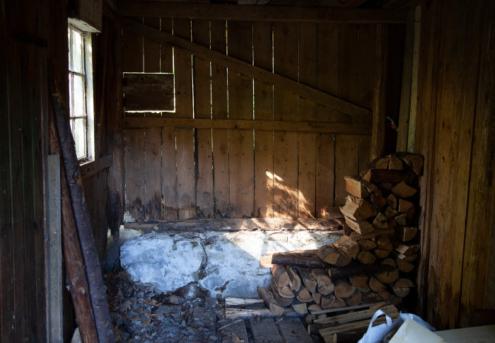 Rymlig vedbod med slitet trägolv och väggar samt en hög staplad ved vid vitfönstrad vägg.