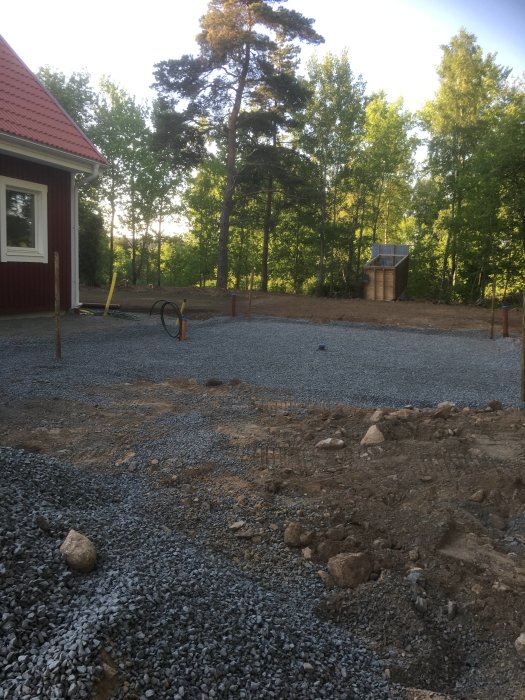 Förberedd byggplats med grus och förgjutna fundament, omgiven av gröna träd och en röd husknut.