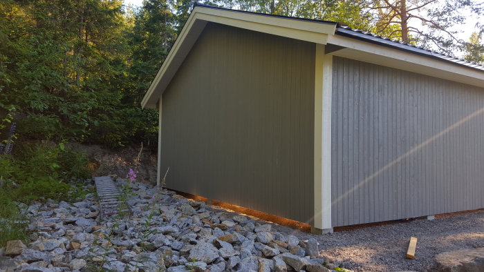 Nybyggd grå carport med vita knutbrädor och takfot, omgiven av skog och grusgrund.