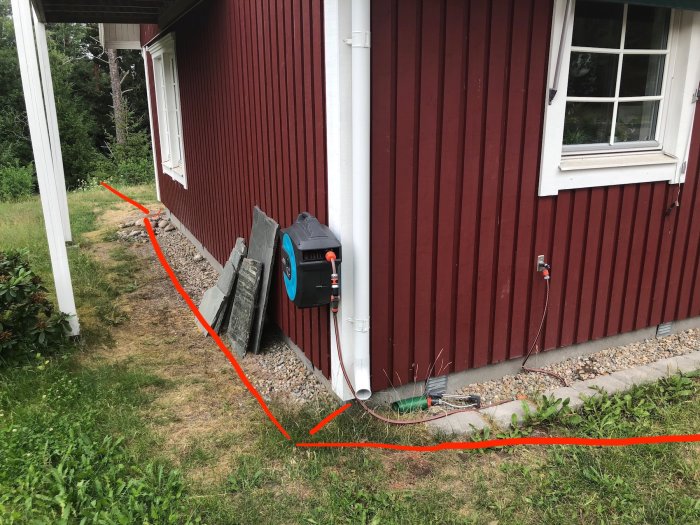 Husröd vägg med stuprör, plan för dräneringsrör markerad med röda linjer på en lantlig tomt.