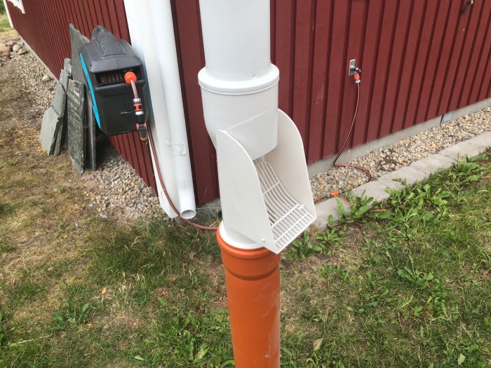 Stupränna med vattenuppsamlare ansluten till orange dräneringsrör utomhus vid röd husvägg.