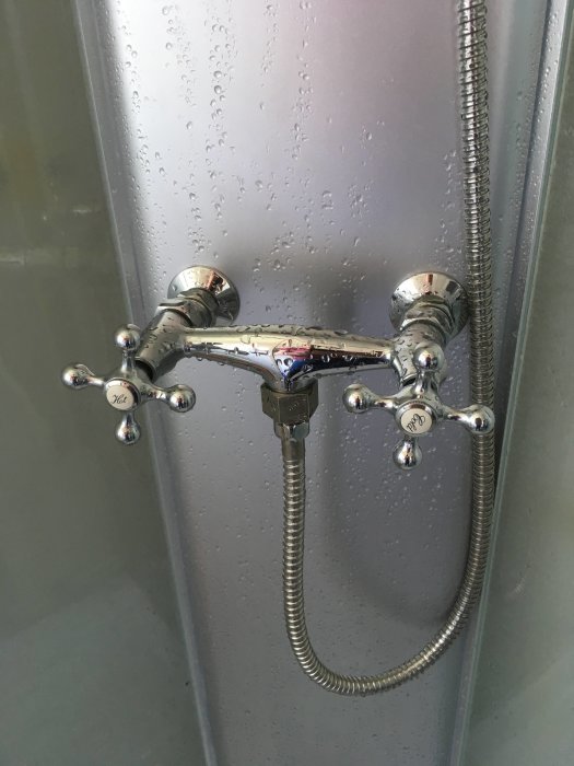 Kromad duschblandare med klassiska kranar och ansluten duschslang på en våt kakelvägg.