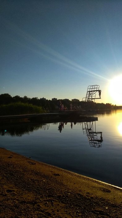 Kvällssol vid en strand med människor på en brygga och reflektioner i vattnet.
