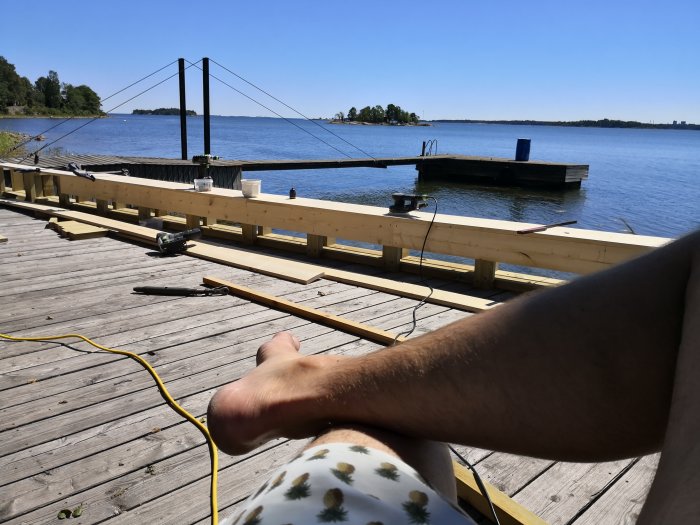 En person som vilar benen på en träbrygga vid sjön, byggmaterial och verktyg i bakgrunden, soligt väder.