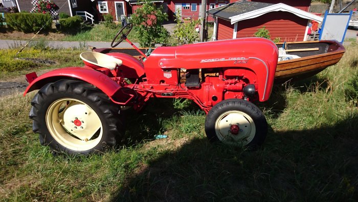 Röd vintage Porsche-Diesel traktor parkerad utomhus i solsken med en båt i bakgrunden.