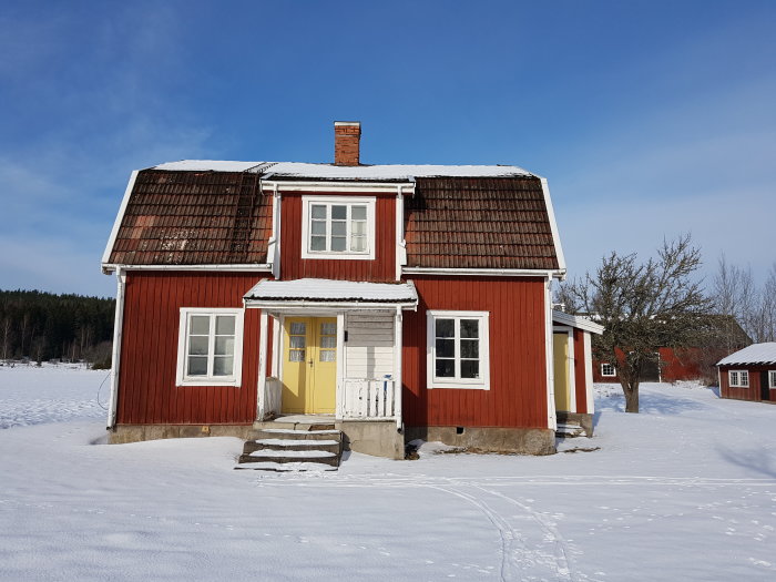 Rött trähus med vita knutar och snötäckt tak i ett vinterlandskap, markerat för rivning.