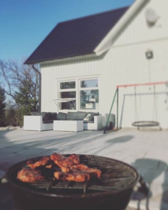 Ej färdigställd uteplats med grå plattor och vita murar, halvfärdigt grillkött i förgrunden, vit husfasad i bakgrunden.
