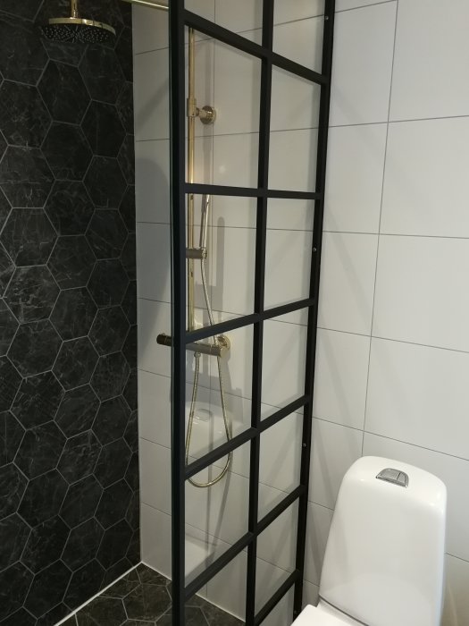Nytt modernt badrum med svart duschvägg, vita och svarta kakelväggar samt guld duschdetaljer.