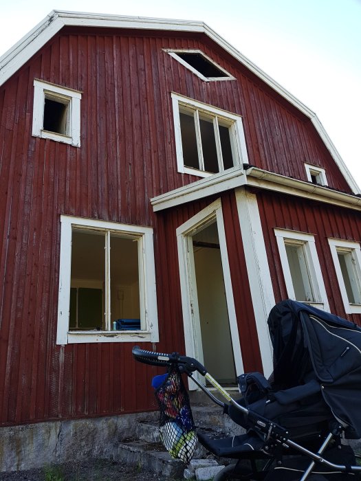 Rödfärgat hus med borttagna fönsterrutor och öppen dörr, redo för rivning, barnvagn framför.