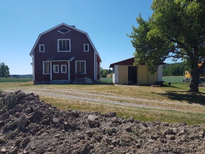 Grävning framför röd lantgård med gul grävmaskin till höger, upphögar med jord i förgrunden.