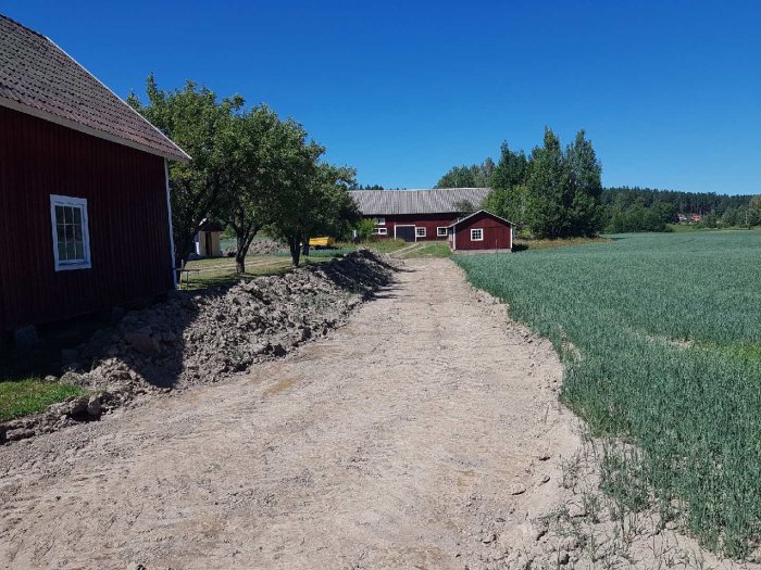 Nygrävd väg omgiven av grönt fält och röda ladugårdar under en klarblå himmel.