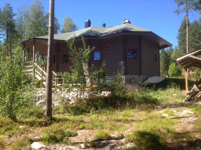 Ett trähus omgivet av sommarvegetation under en klarblå himmel.