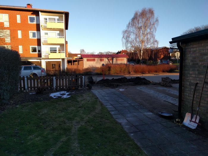 Långsmal trädgård framför radhus med parkerad bil, trottoar, gräsmatta och en hög med jord; planering för renovering.