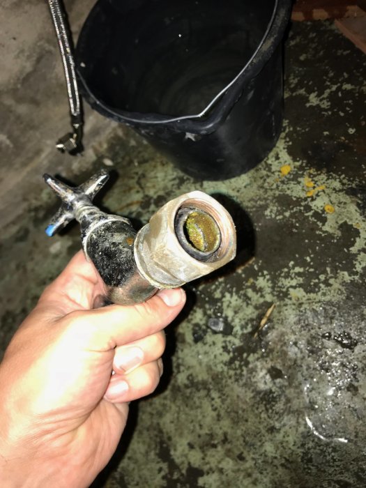 Hand håller ett gammalt rör med gängor nära kamera, med en hink och betonggolv i bakgrunden.