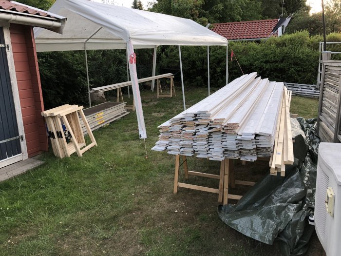 Trädgård med uppsatt målartält och hög med nyinköpt panelbredd och byggmaterial i förgrunden.