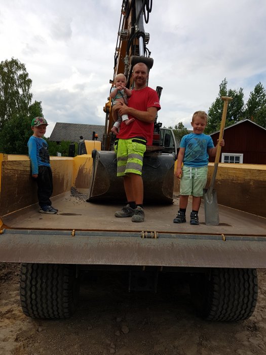 Man och tre barn står framför grävmaskin, ett barn håller i en spade och ett annat barn hålls i famnen.