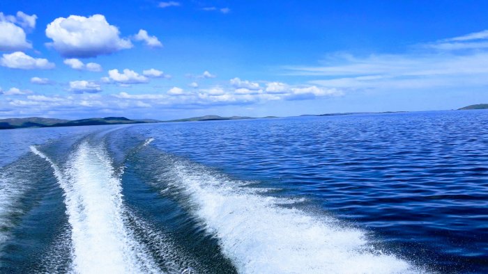 Vy över hav med slät vattenyta och vita molntussar mot blå himmel, vågskum från båt.