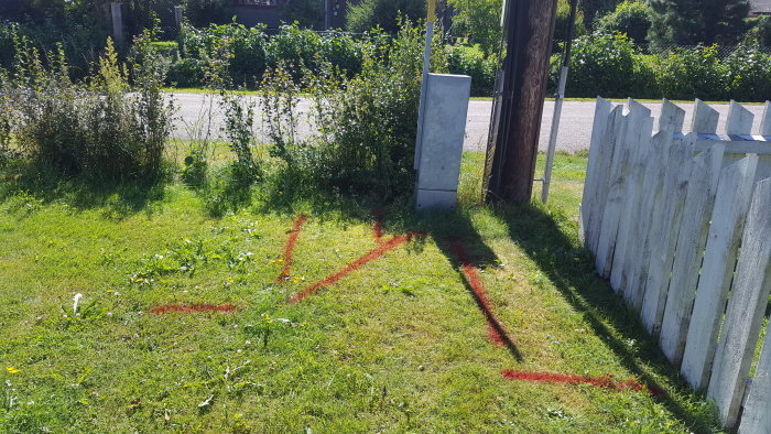 Tomtgräns markerad med röd sprayfärg nära staket, buskar och elskåp.