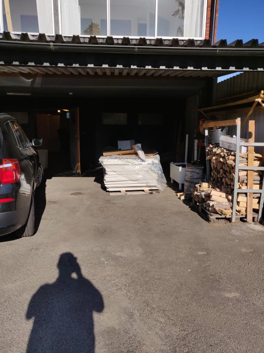 Gipsskivor staplade utomhus vid en garageuppfart med bil och vedhög i bakgrunden.