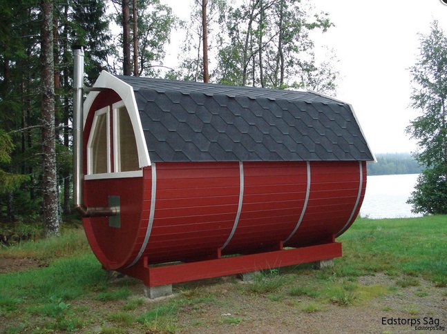 Röd bastutunna av trä vid en sjö med svart tak och vita detaljer, i en skogsmiljö.