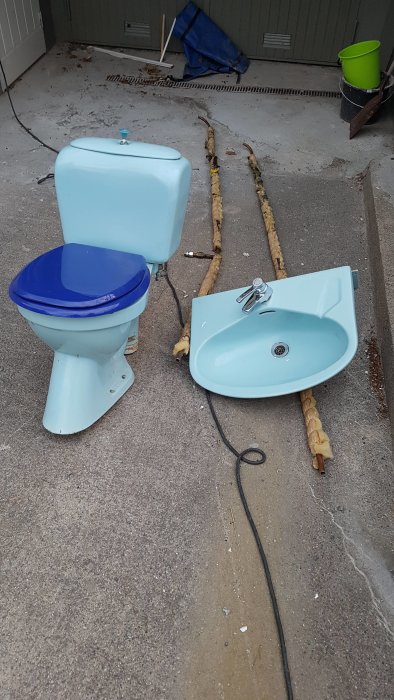 Blått toalettstol och handfat med kran i begagnat skick på betonggolv utomhus.