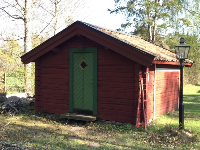 Röd trästuga med grön dörr och rutor samt ett gammalt gatljus, omgiven av natur, potentiellt tjärad doft.