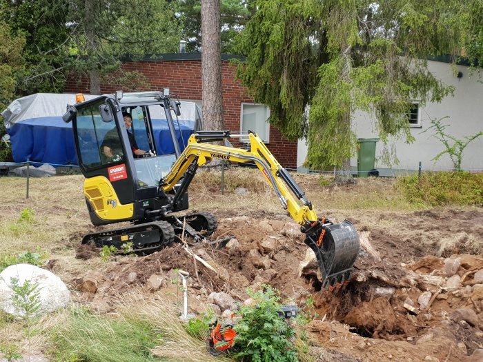 Minigrävare i arbete med att gräva upp en tomt nära en byggnad under sommaren.